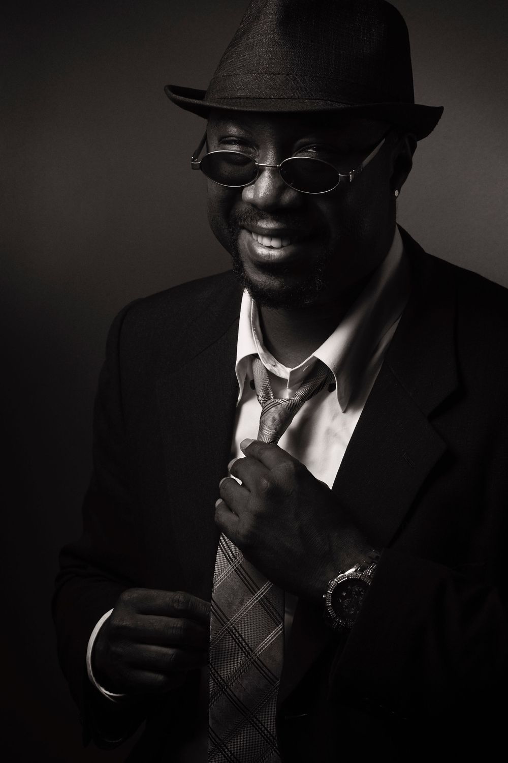 Black and white portrait of a smiling person, Denis, wearing a fedora hat, glasses, suit and tie in dramatic lighting photographed by Evan