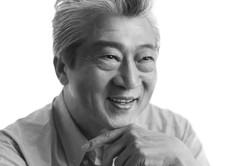 A black and white portrait of Hiroyuki Ishii in casual clothing with a warm smile against a light background photographed by Evan Will