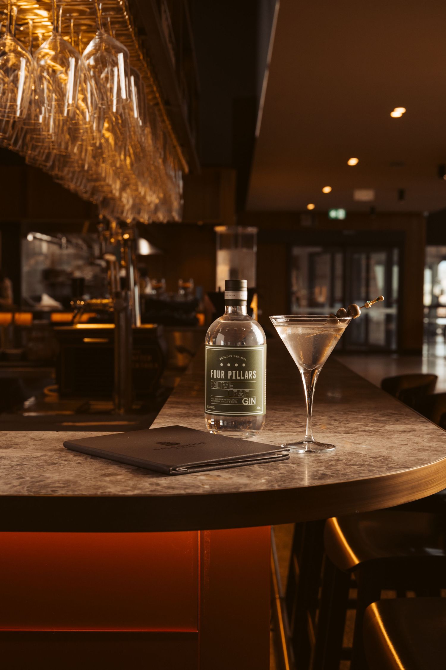 A sleek bar setting showcasing a bottle of Four Pillars Gin beside a martini glass with garnishes, illuminated by warm lighting and hanging