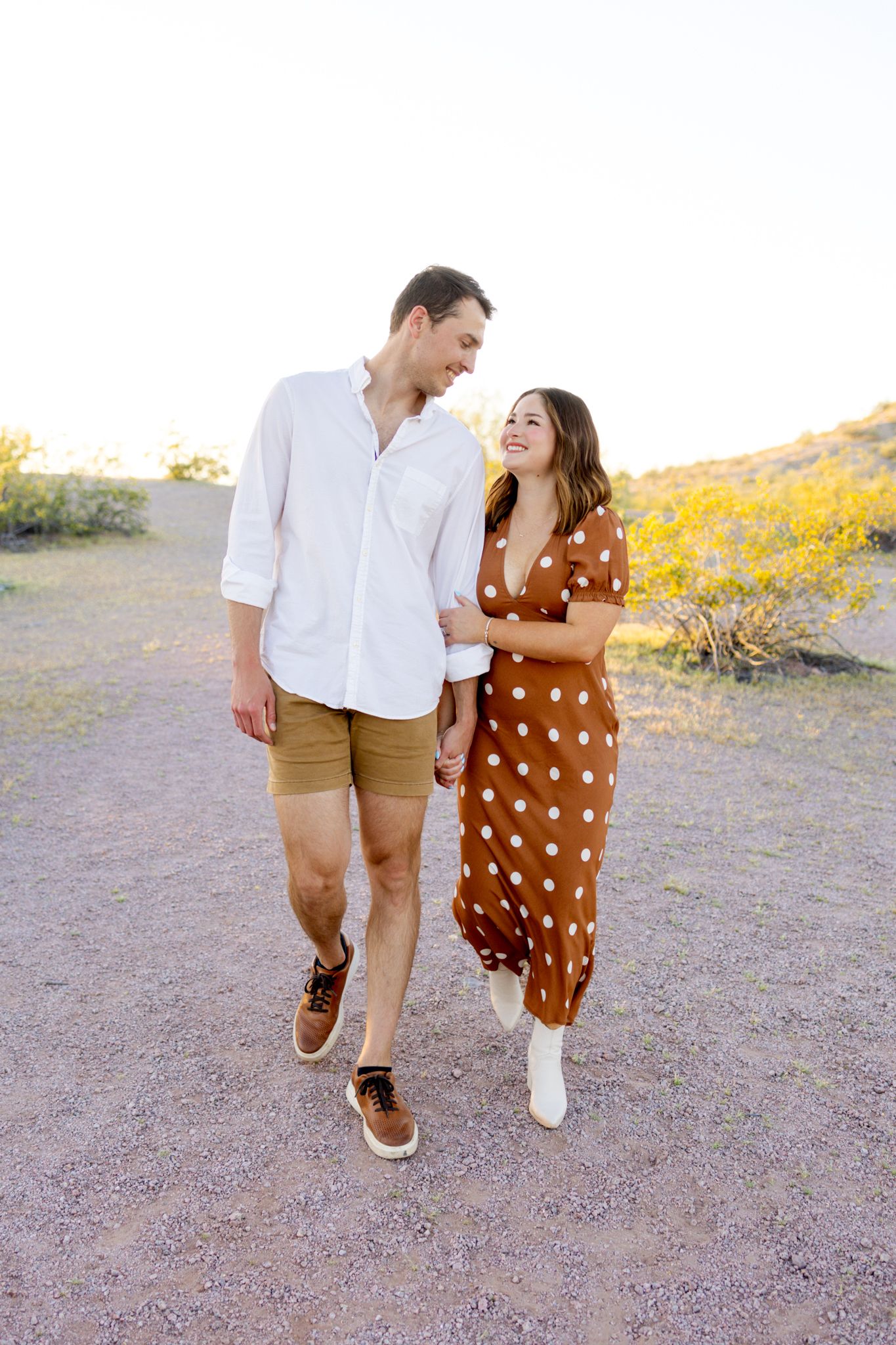 A couple walks together and laughs outdoors, wearing casual outfits with her in a brown polka dot dress and him in khaki shorts.