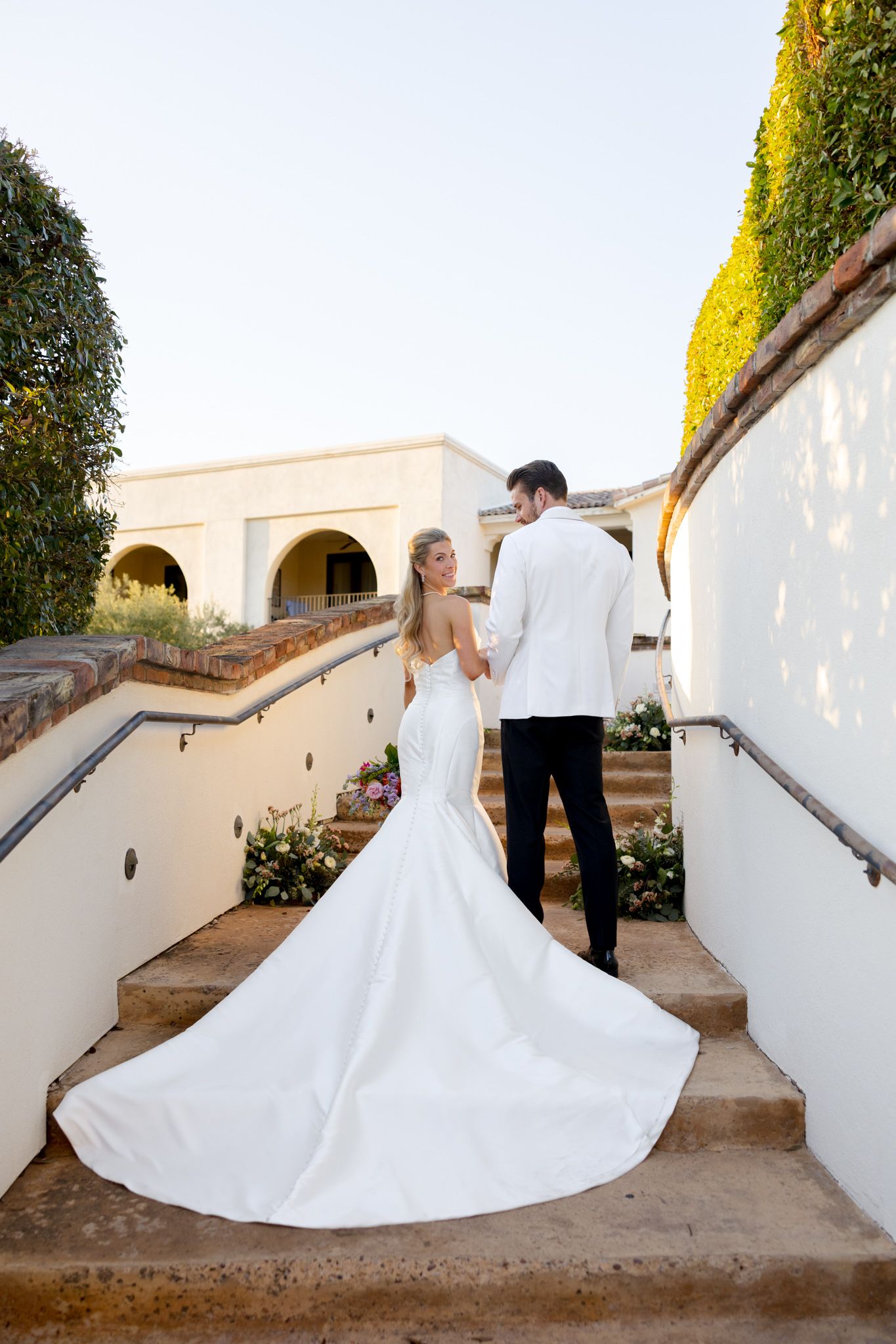 A bride in a flowing white mermaid gown and groom in a tuxedo stand on stone steps at an elegant venue with arched architecture.