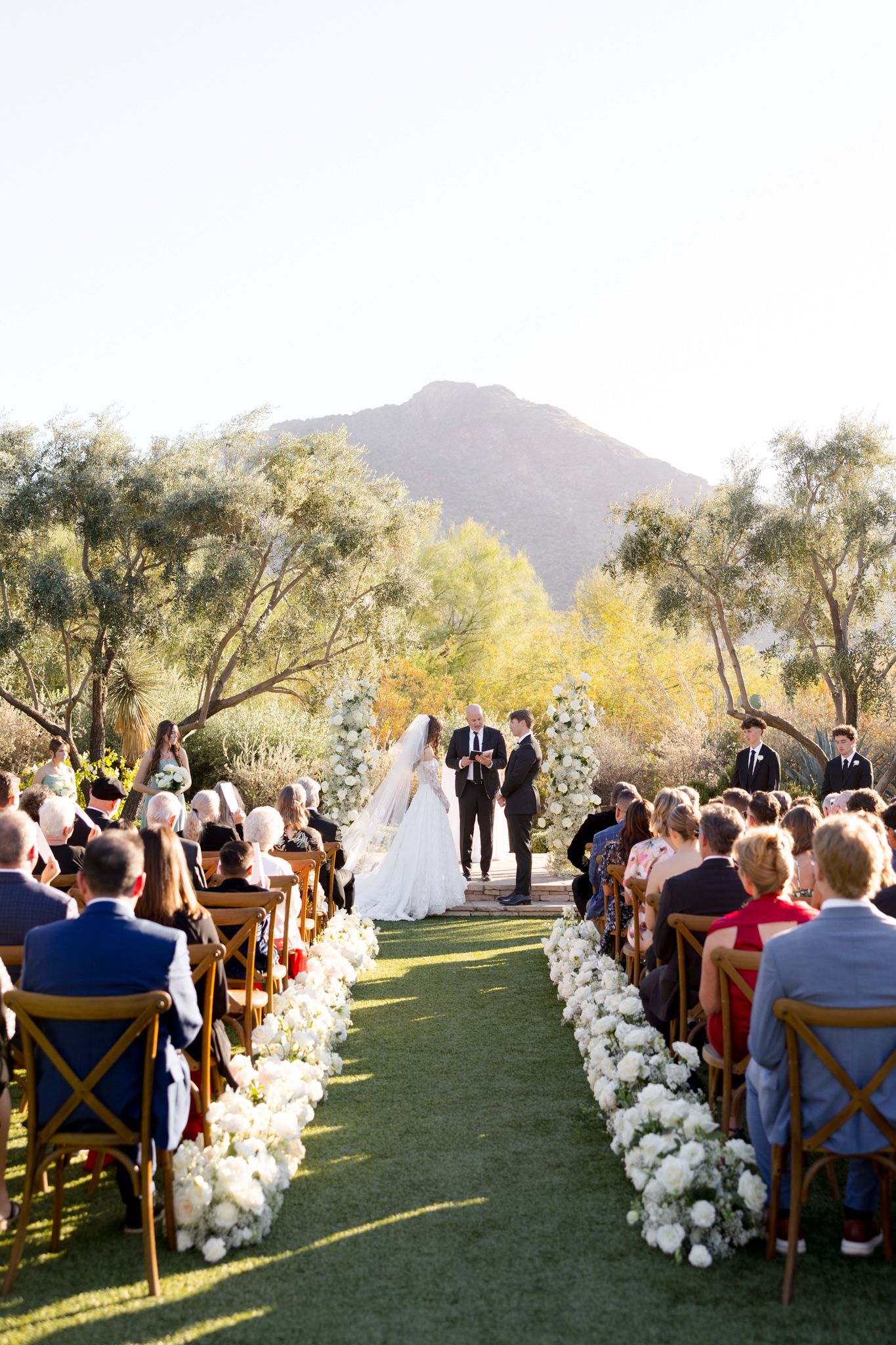 Outdoor desert wedding ceremony set against mountain backdrop with white floral aisle decorations and wooden cross-back chairs.