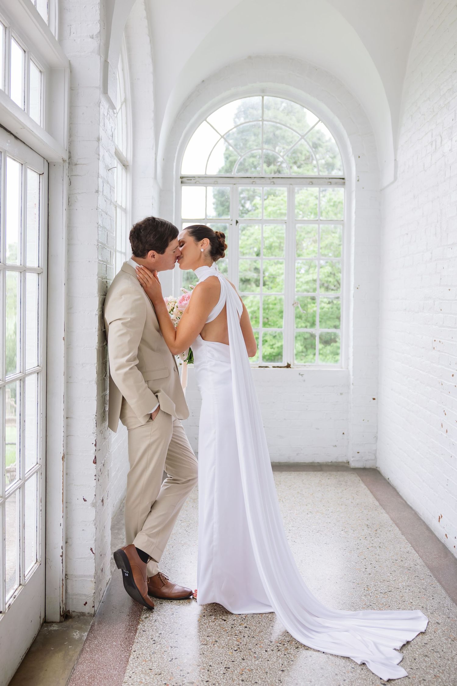 A couple shares a romantic moment in an elegant white hallway with arched windows and natural light.
