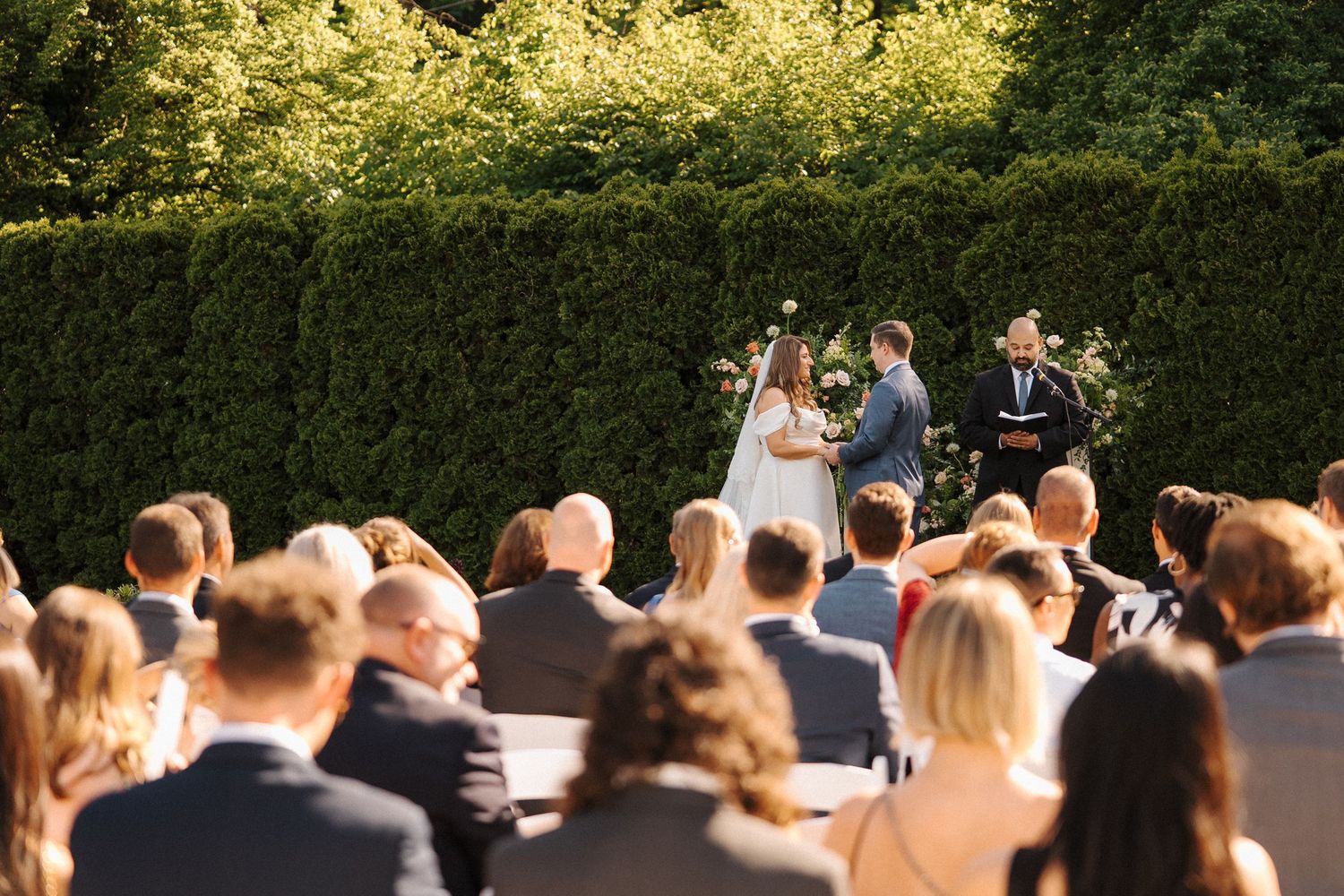 Outdoor garden wedding ceremony with guests seated in rows facing couple and officiant by manicured hedge wall.