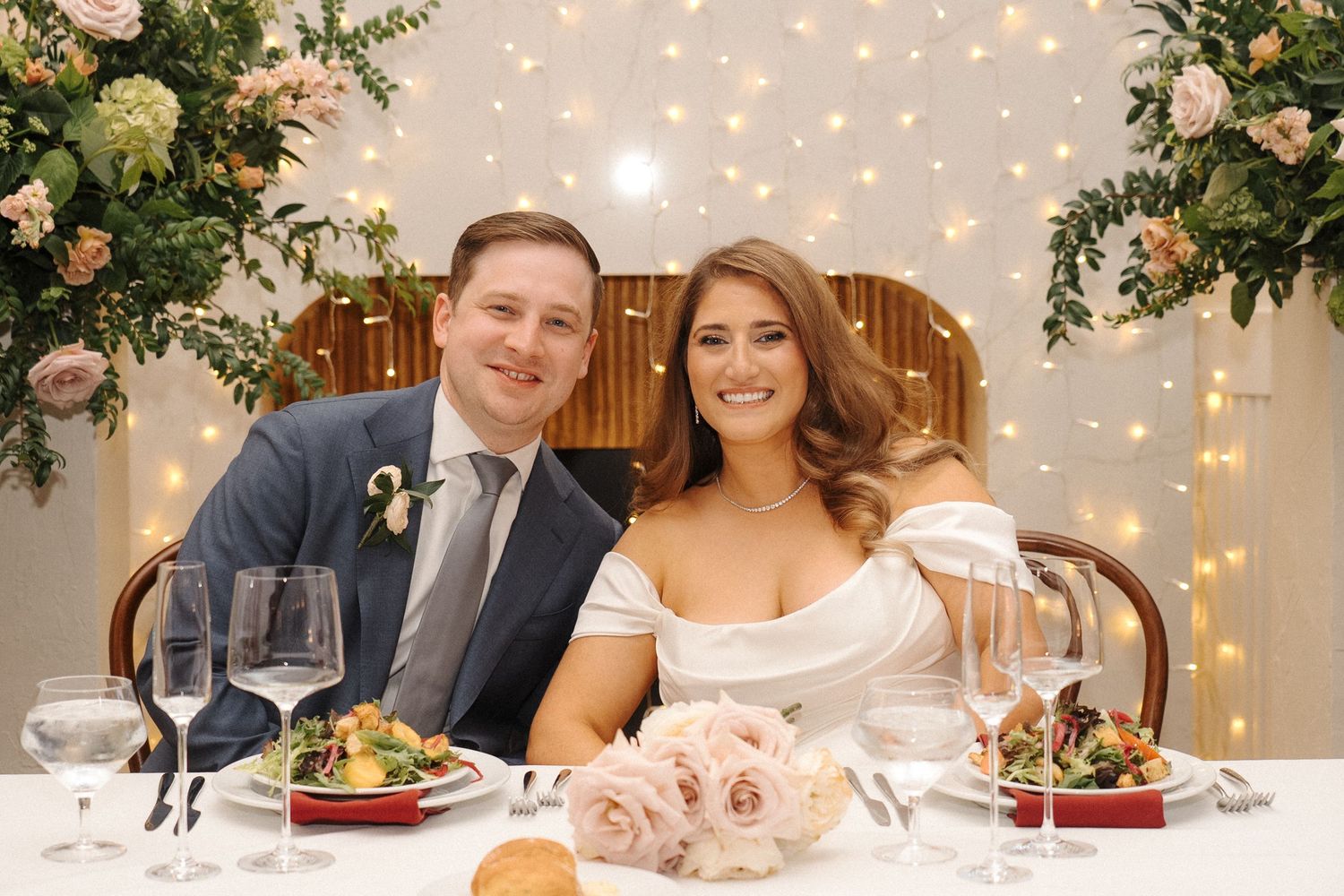 Table setting at an elegant wedding reception with string lights, floral arrangements and guests seated for dinner service.