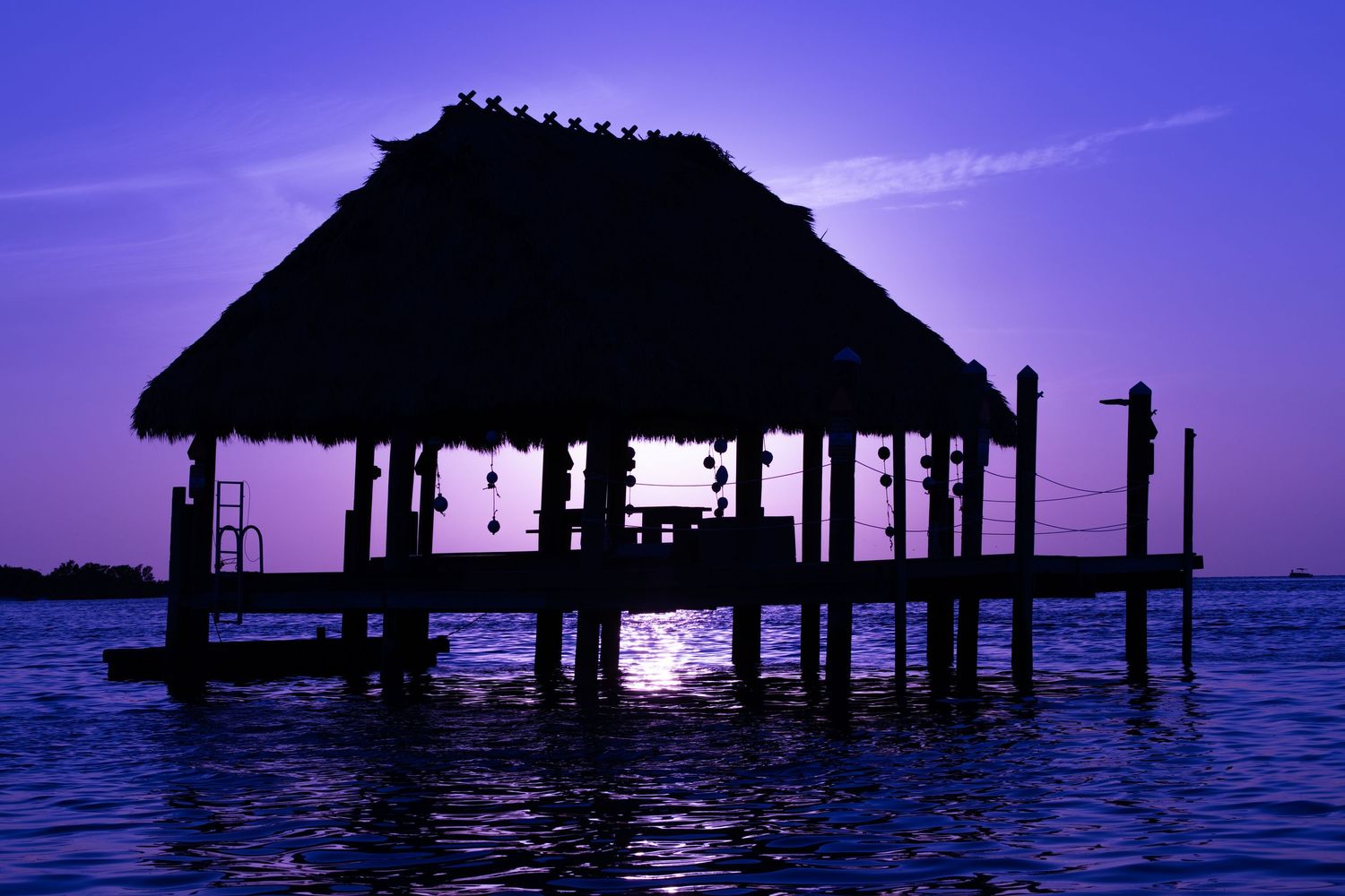 Silhouette of a thatched-roof pier hut over calm blue waters at sunset with purple and pink sky.