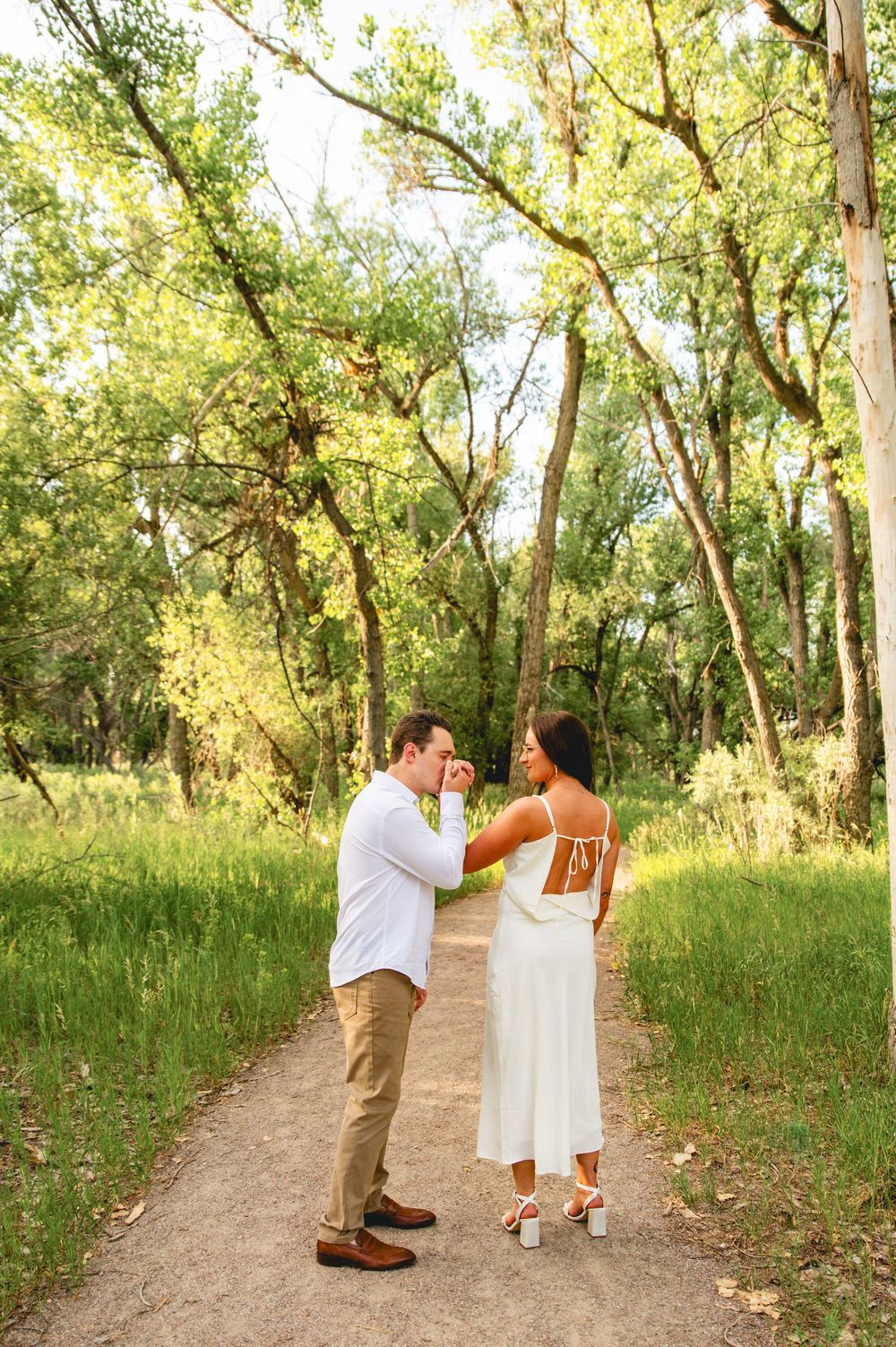 A couple shares a romantic dance moment on a tree-lined dirt path as warm sunlight filters through the leaves.