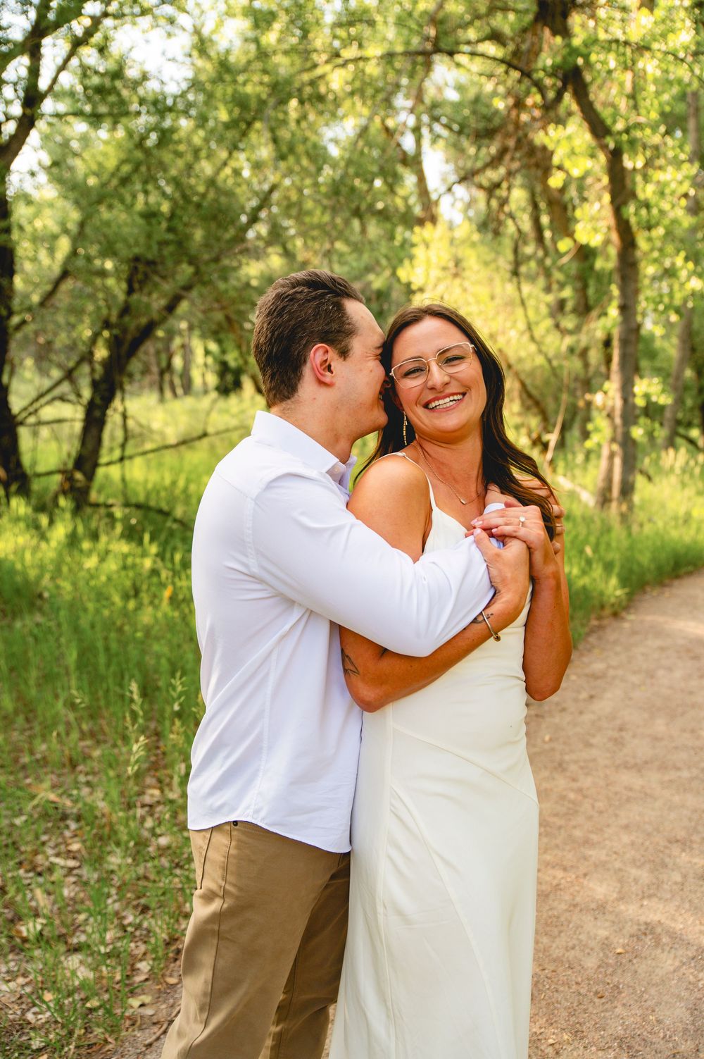 A couple shares a romantic moment while laughing together on a sunlit dirt path surrounded by lush green trees.