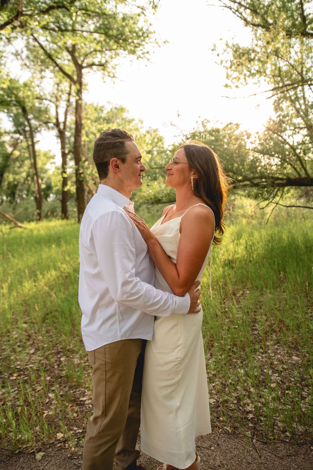 A couple embraces during sunset in a wooded park setting while wearing white and casual summer attire.