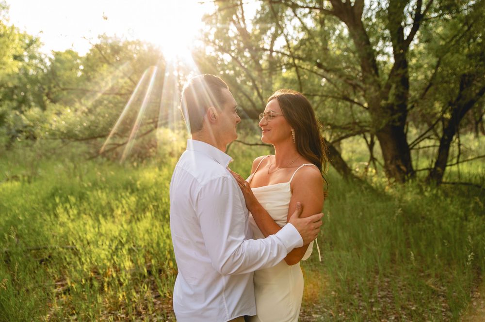 A couple embraces in golden sunset light while standing in a lush green meadow surrounded by trees.