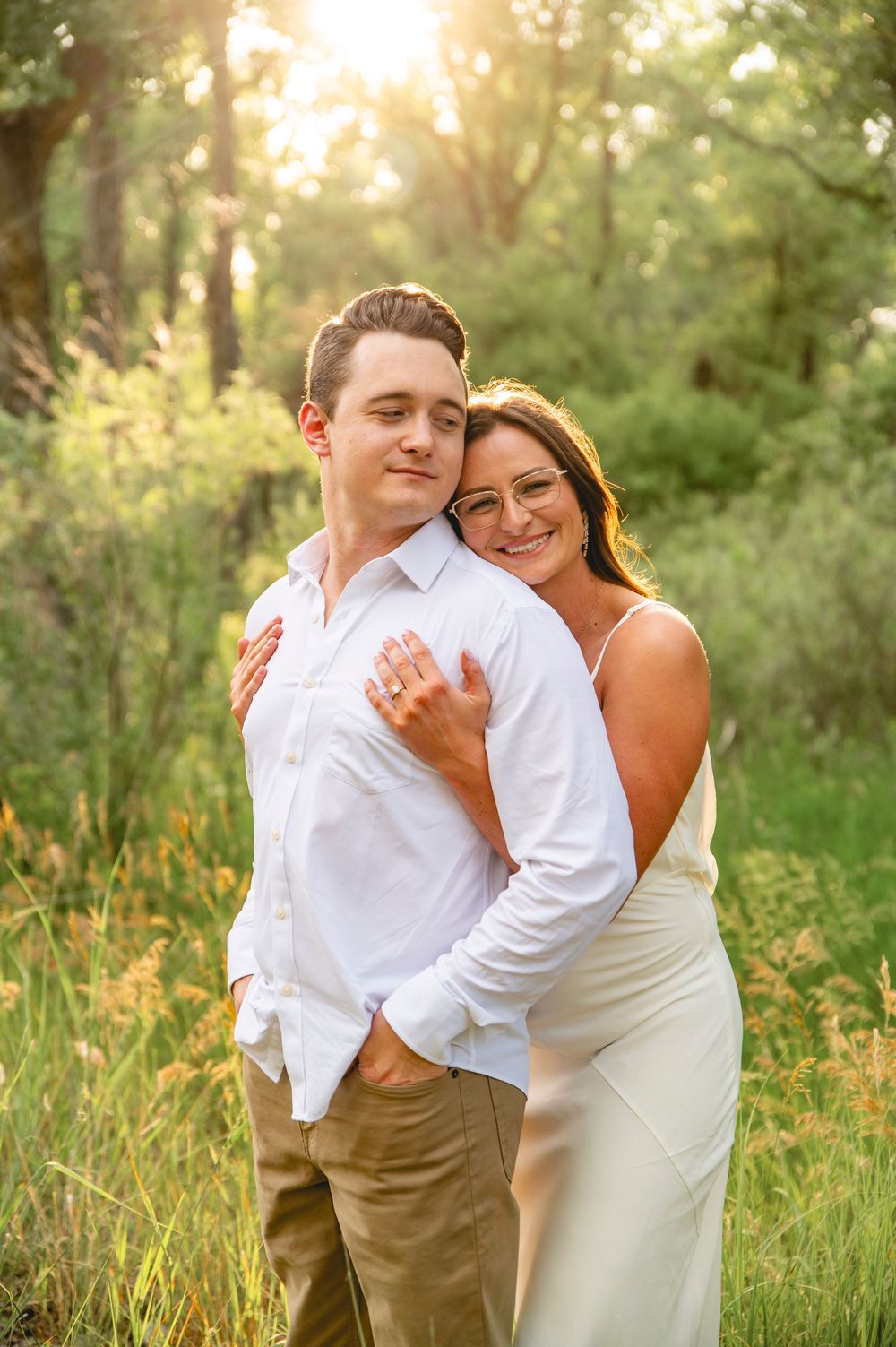 A couple in wedding attire embraces while standing in a sunlit meadow with tall grass and trees at golden hour.