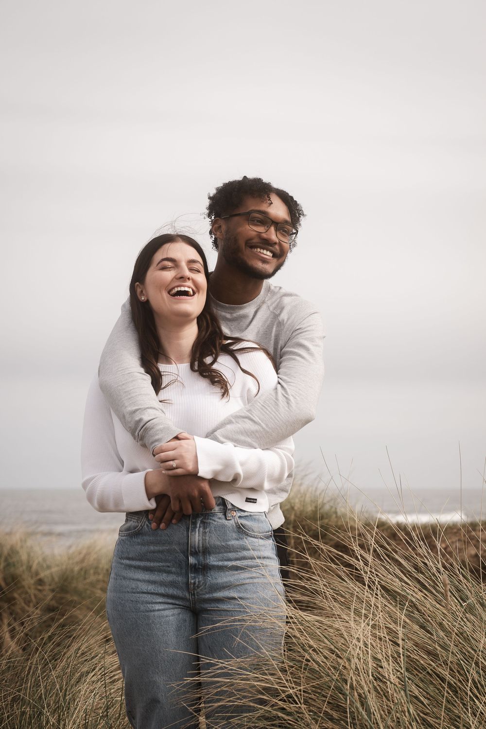 Happy couple embracing and laughing together on a misty beach with tall grass in the foreground.