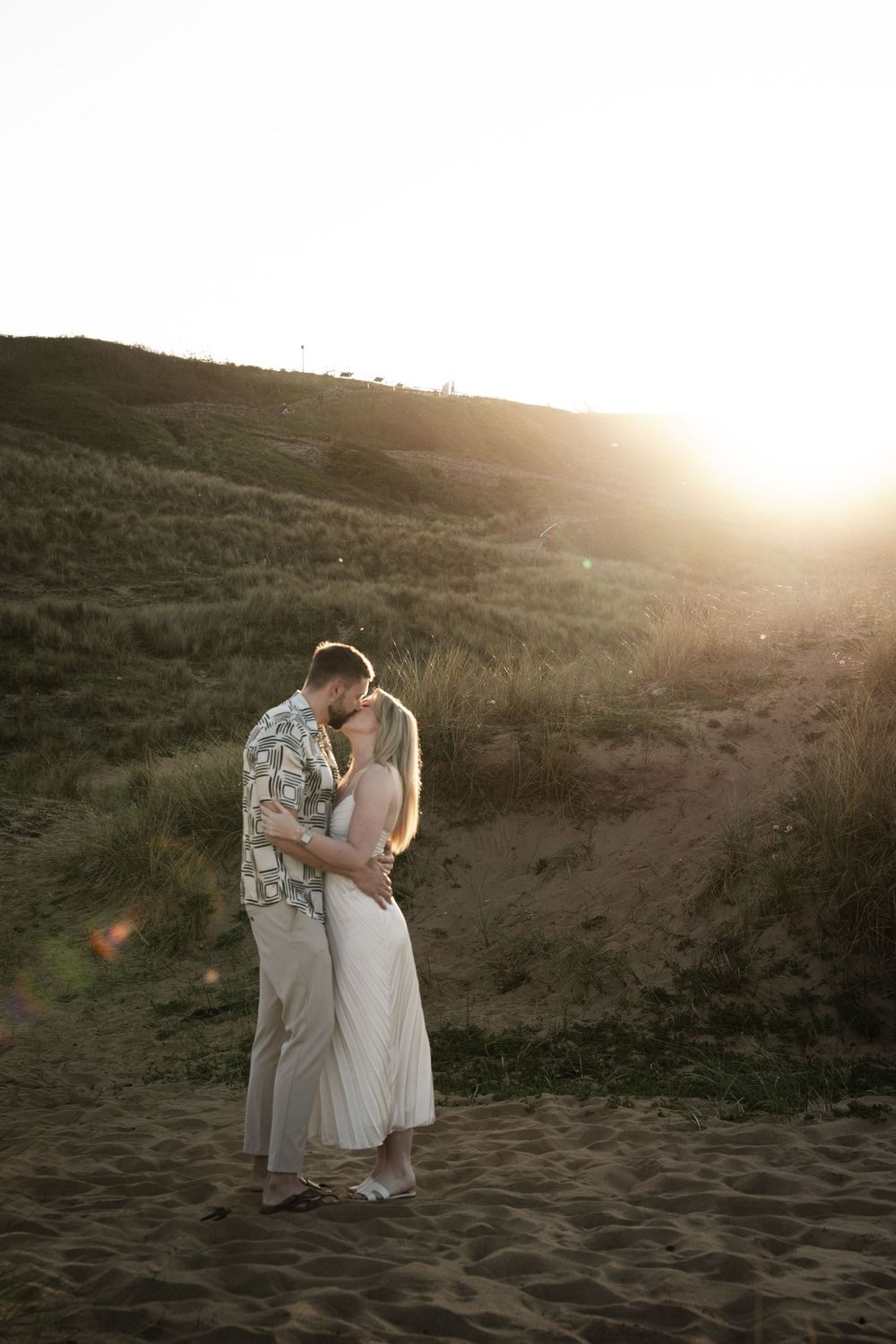 A romantic couple embraces on a sandy beach at sunset with golden sunlight streaming behind them.
