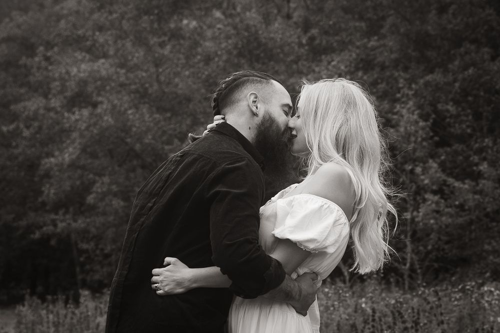 A romantic black and white portrait of a couple embracing outdoors against a wooded backdrop.