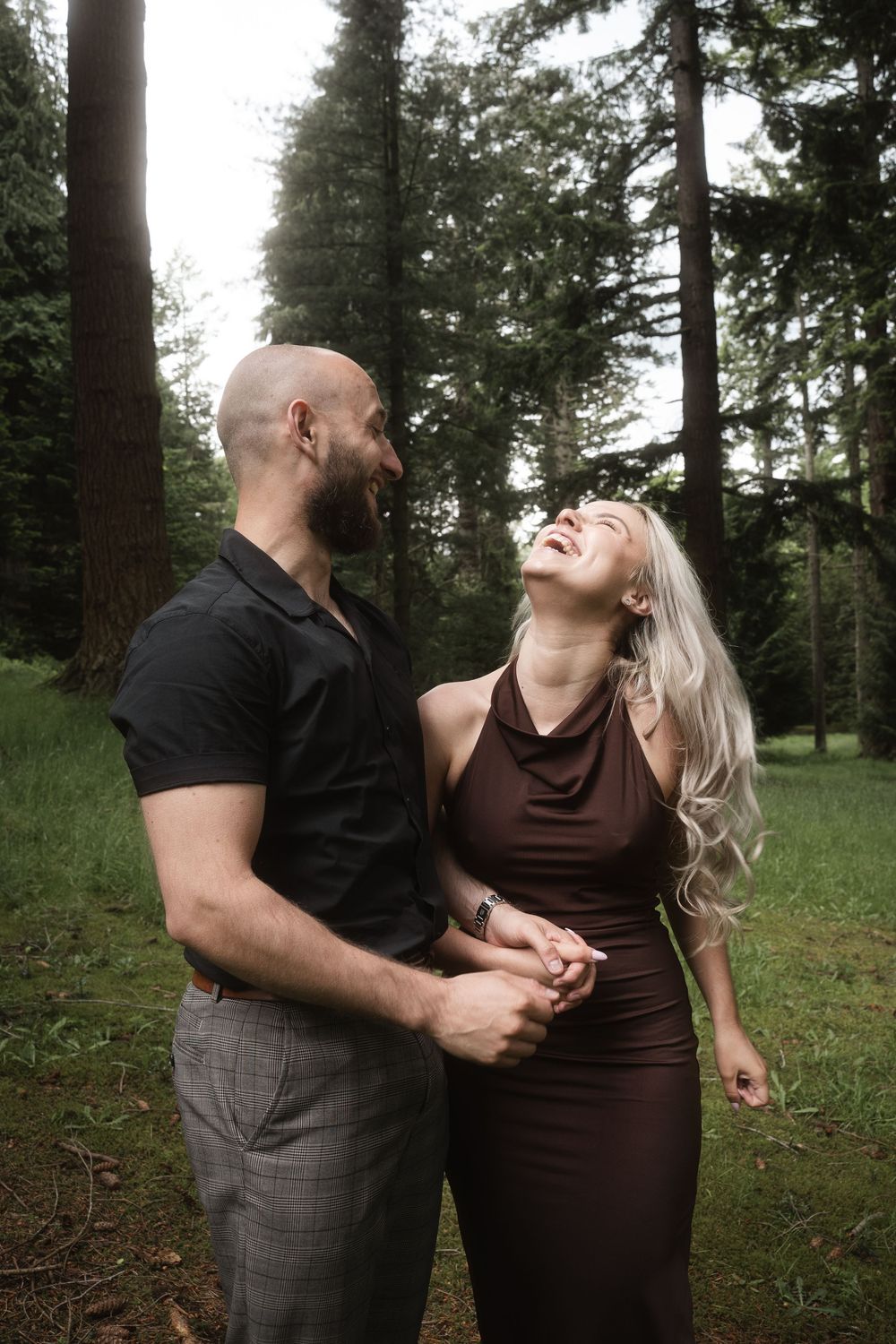 A couple shares joyful laughter together while embracing in a wooded outdoor setting.