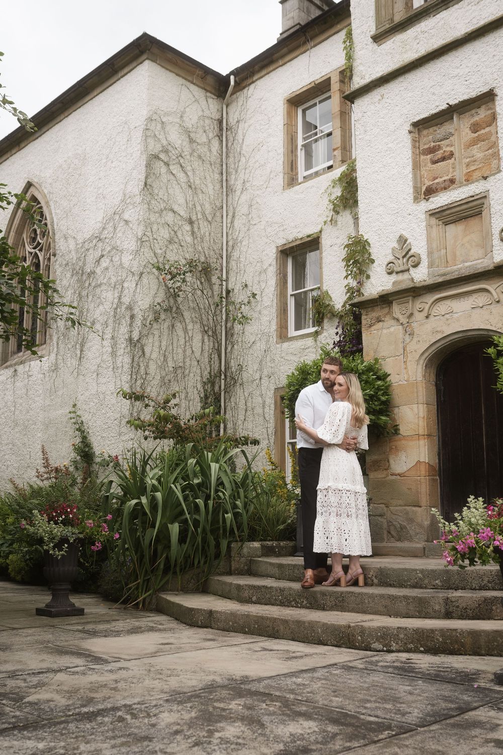A couple embraces on stone steps outside a white stucco building with climbing vines and greenery.