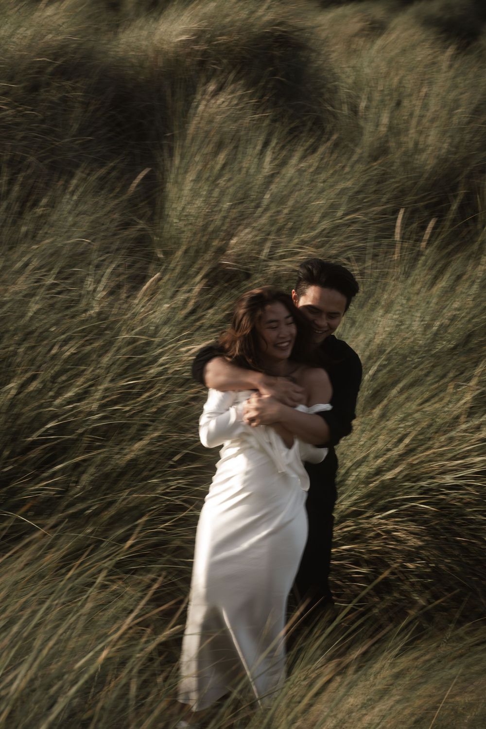 A romantic couple embraces in tall wild grasses, with the bride wearing a flowing white off-shoulder dress at sunset.