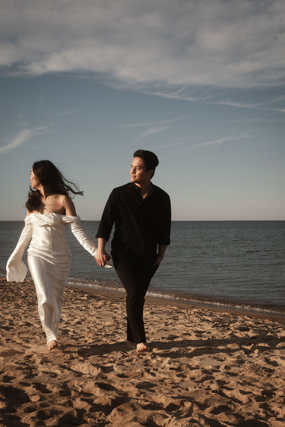 A couple in black and white outfits walk along a sandy beach with dramatic cloudy skies over the ocean.