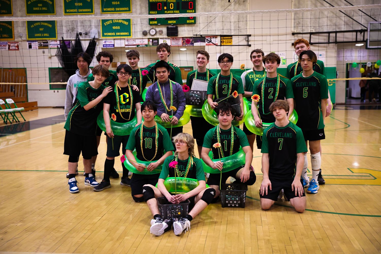 A volleyball team wearing green jerseys sits together on a wooden gymnasium floor for a group photo.