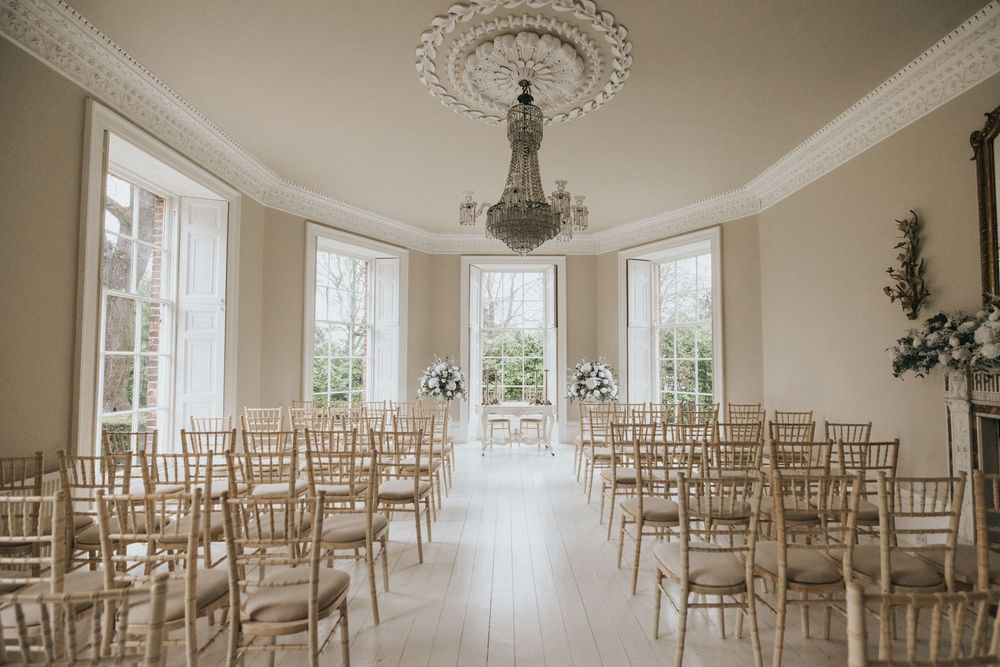 Bright white wedding venue with ornate ceiling details and rows of wooden Chiavari chairs arranged for a ceremony.