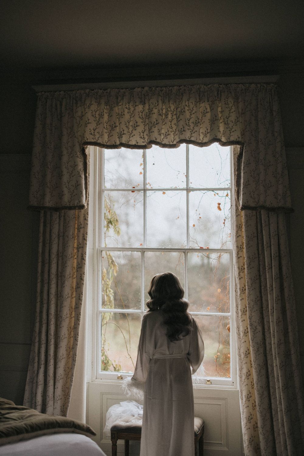 A silhouetted figure in a white dress stands at a tall window with lace curtains gazing out at a misty morning view.