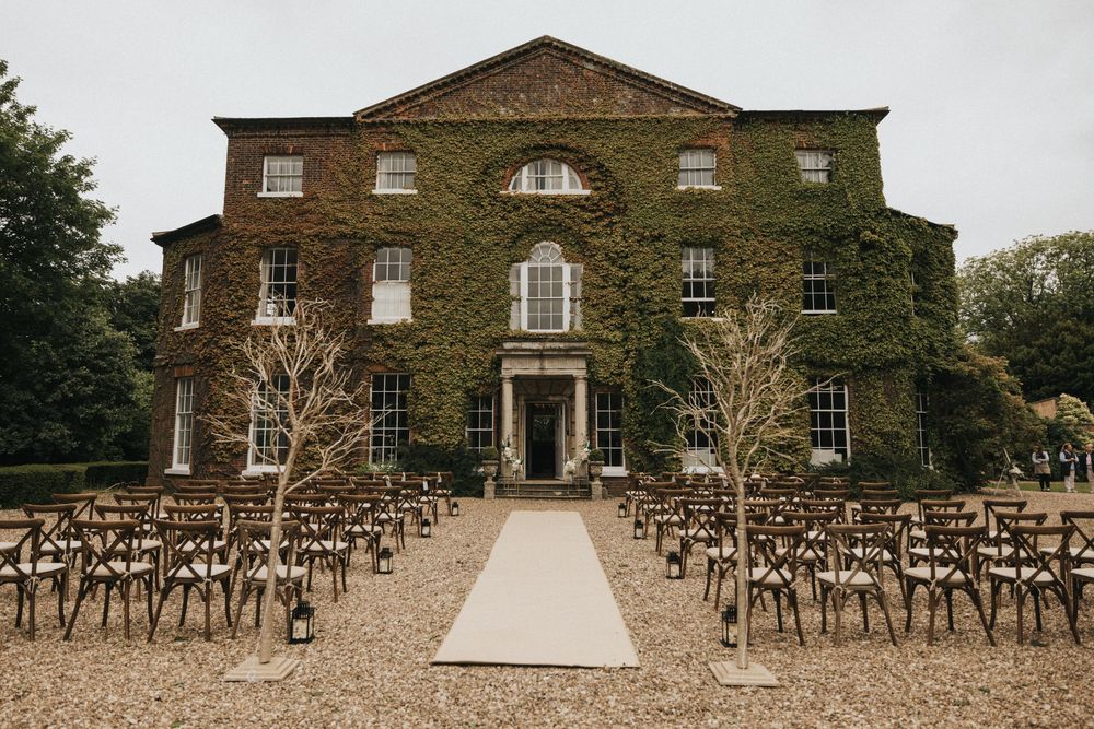 A panoramic view of an elegant stone mansion with rows of wooden chairs set up for an outdoor wedding ceremony.
