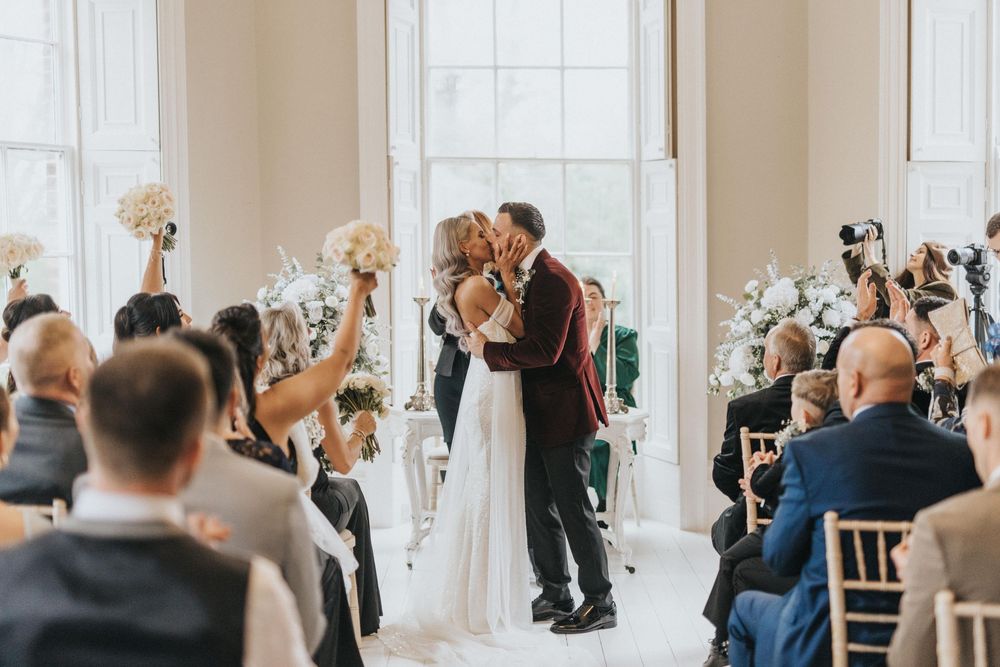 A couple shares their first kiss as newlyweds at a beautiful indoor ceremony with white floral decorations.