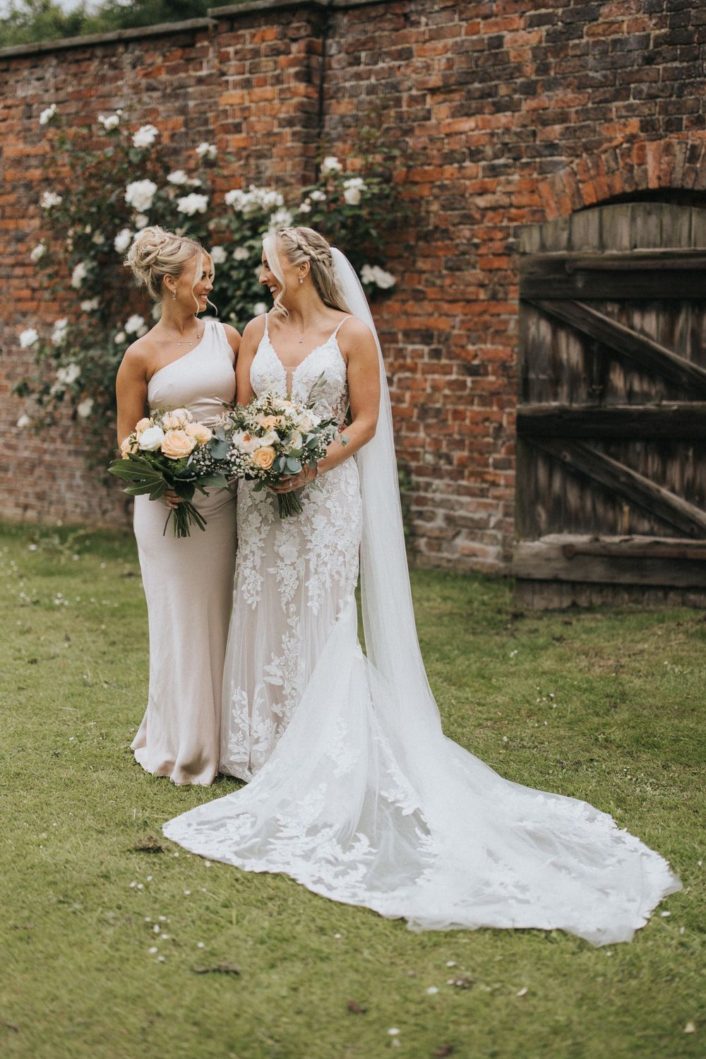 A bride and bridesmaid holding bouquets pose together against a rustic brick wall with white climbing roses.