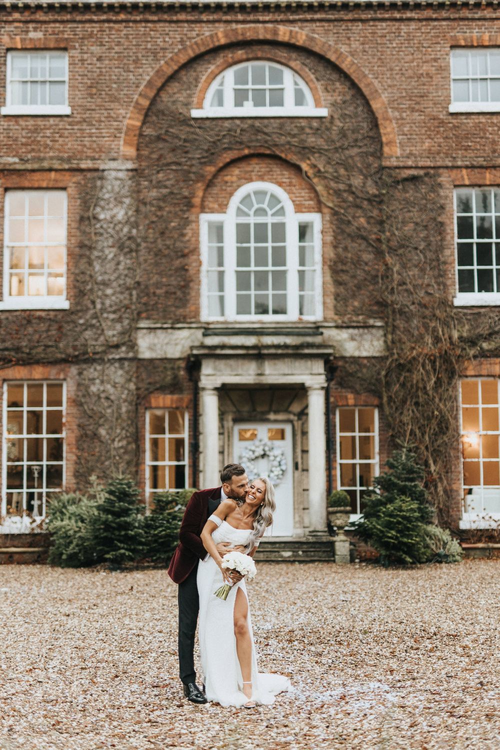 A romantic wedding couple embraces in front of a grand Georgian brick manor house with ivy-covered walls.