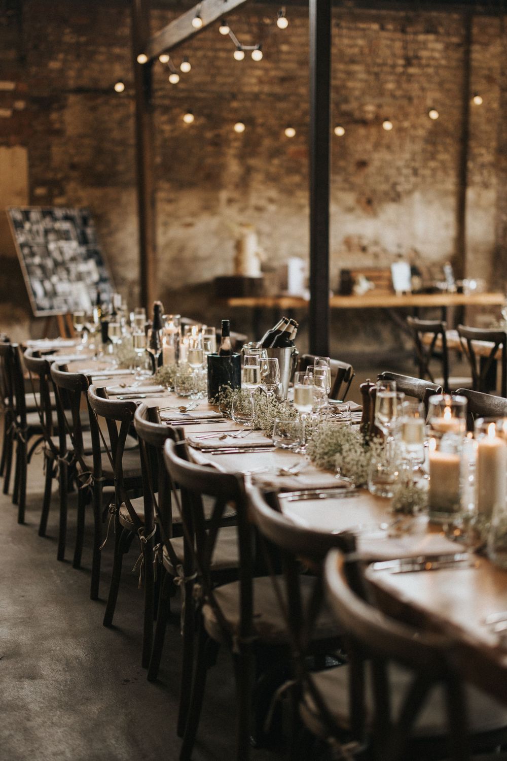 Rustic wedding reception table with dark wooden chairs, string lights, and baby's breath centerpieces in an industrial venue.