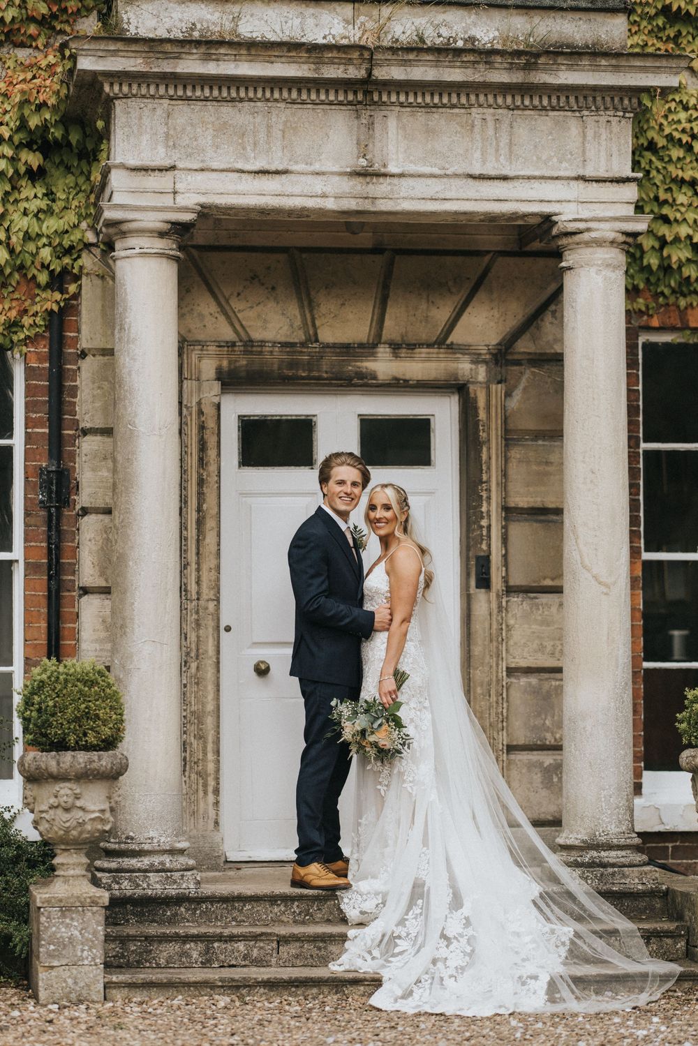 A newly married couple poses elegantly on stone steps in front of a classic columned entryway with ivy-covered walls.