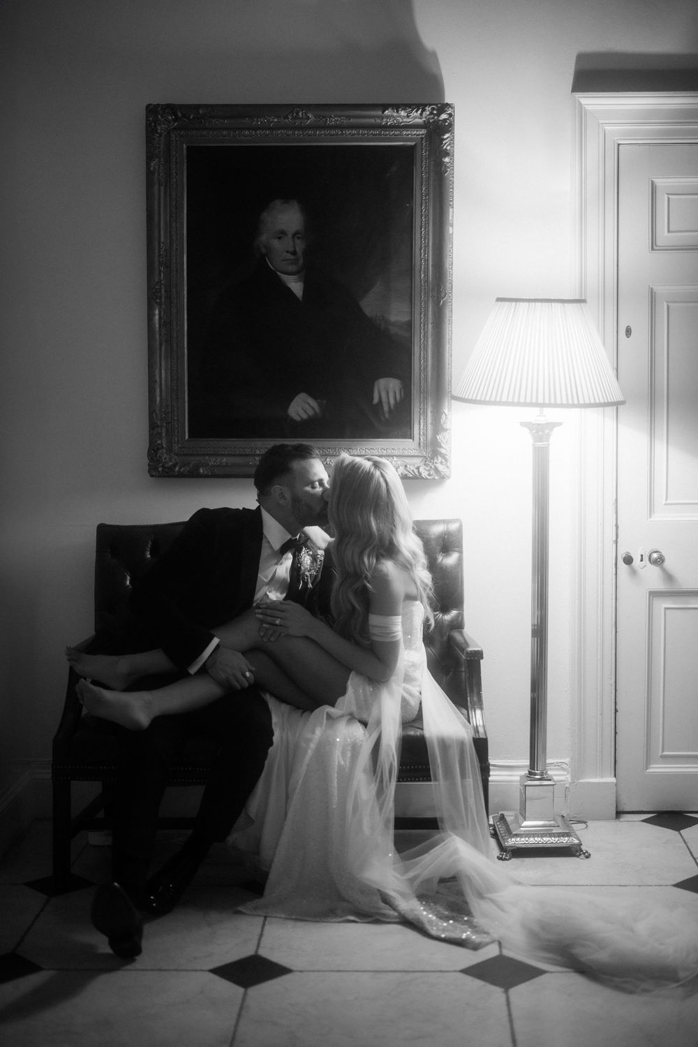 Bride and groom share an intimate moment beneath a vintage portrait in a black and white photograph.