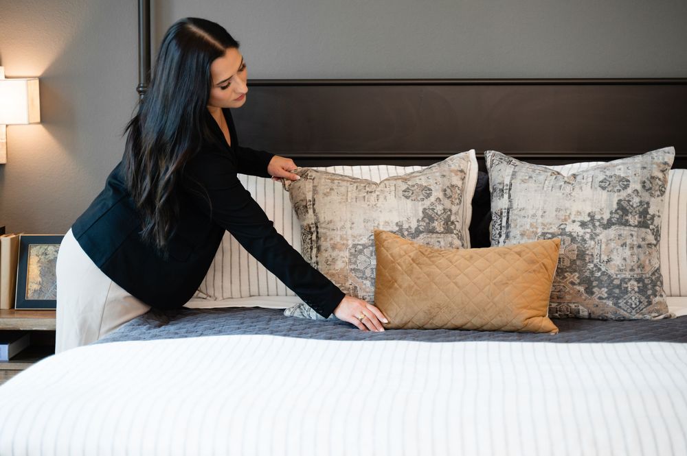 Person making a bed with decorative gray and tan pillows in an upscale bedroom setting.