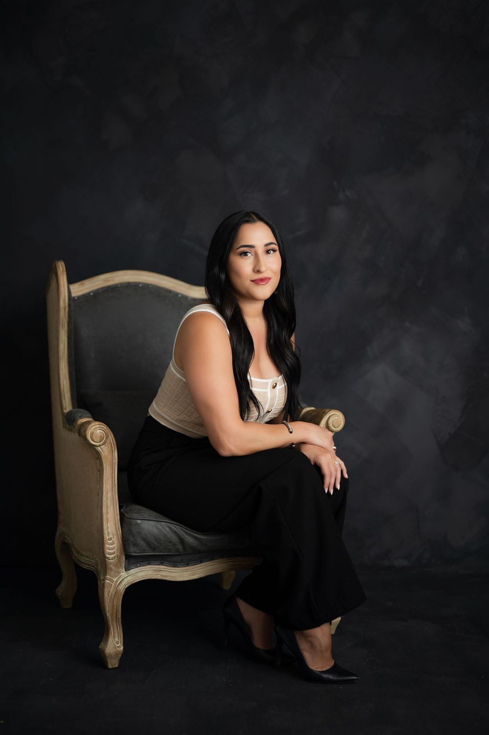 Professional studio portrait of someone seated in an elegant beige chair against a dark dramatic background.