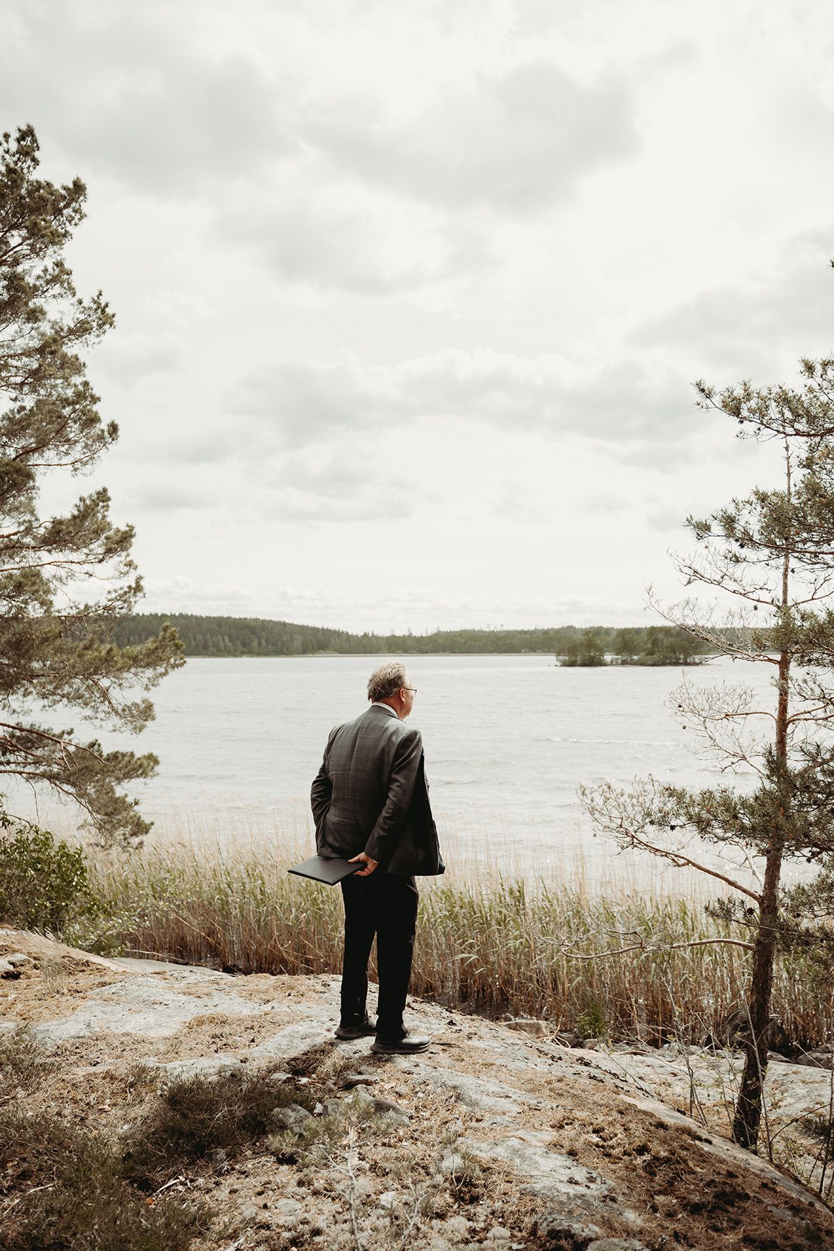 Vigselförrättare står på en klippa med händerna bakom ryggen och blickar ut över vattnet vid vigselplatsen i Sörmland. Fotograf Lets Bloom