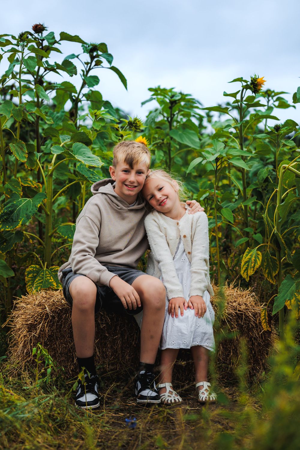Two children sitting together on hay bale in sunflower garden.