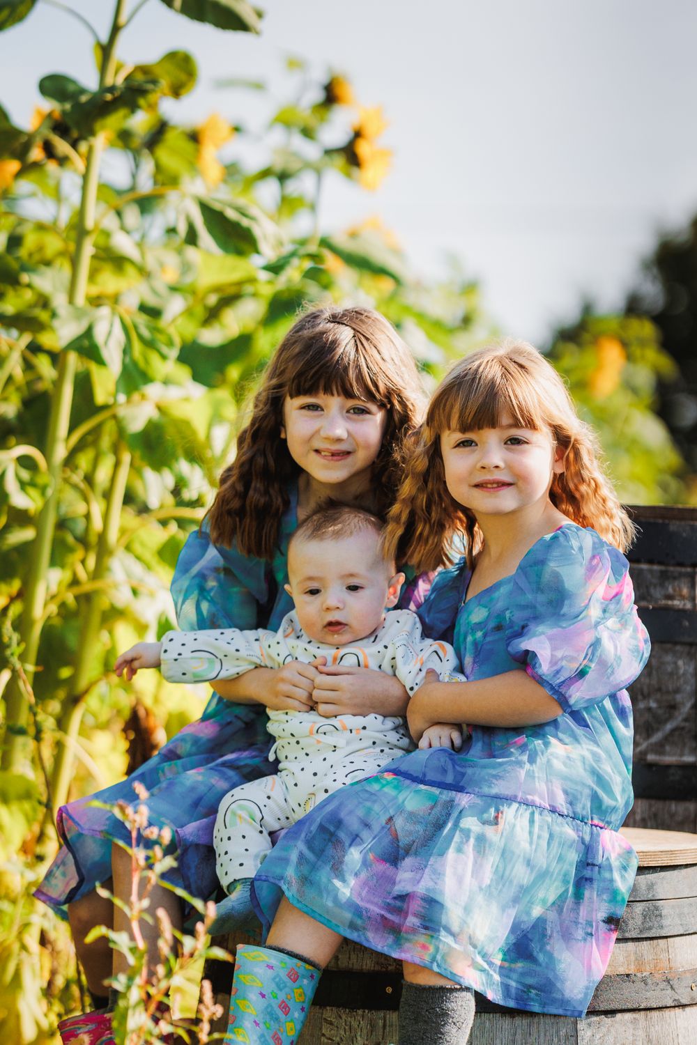 Three siblings in blue tie-dye dresses sitting on wooden bench among sunflowers.
