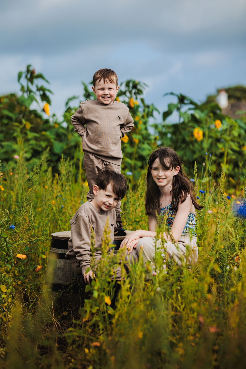 People enjoying sunny day among tall sunflowers and wooden barrels in field.