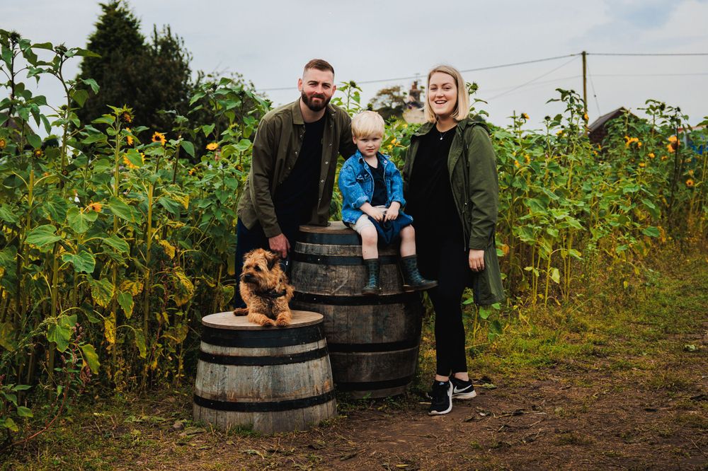 A family portrait with their dog posed on rustic barrels in a sunflower field.