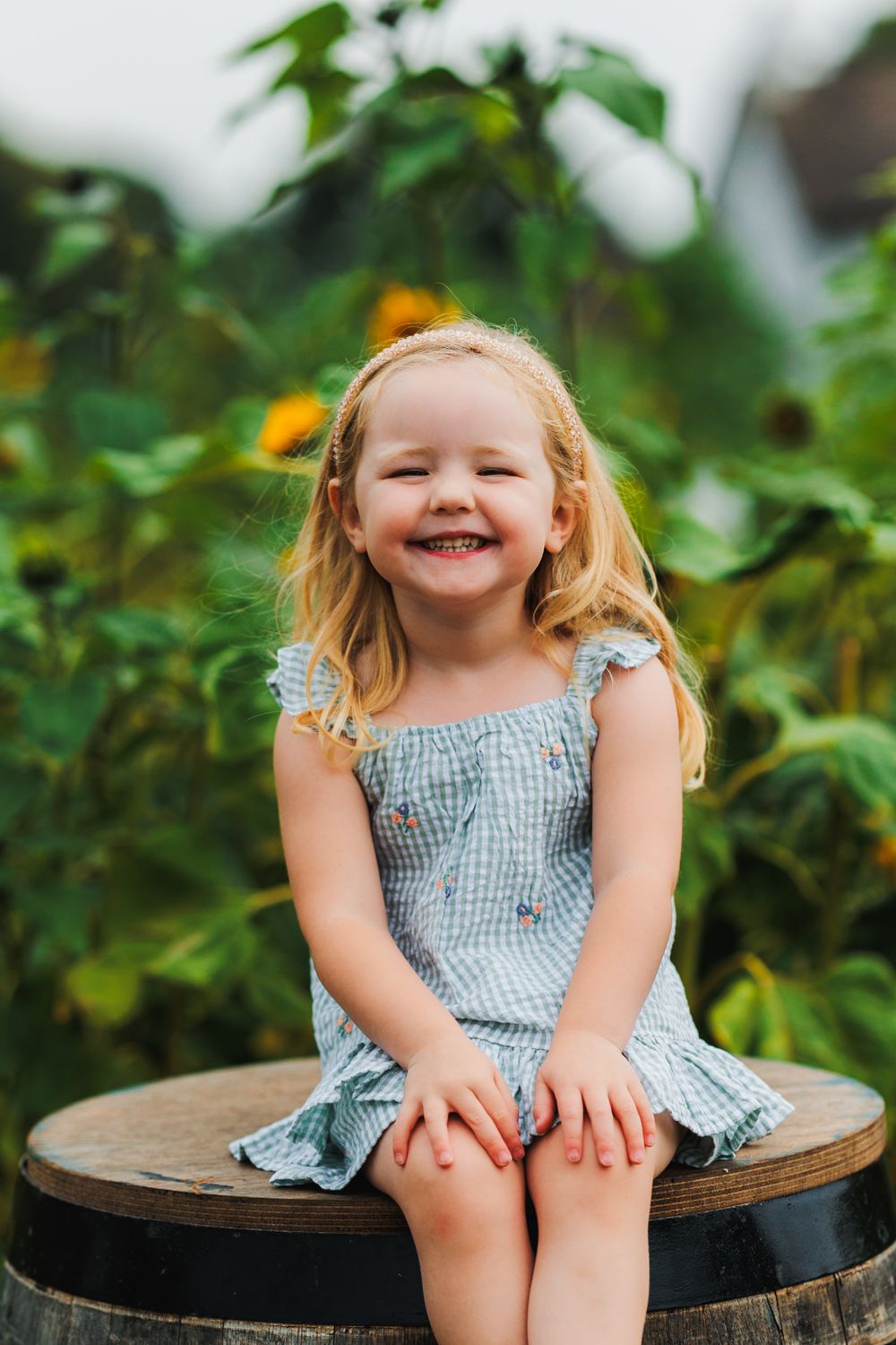 Young child sitting on wooden barrel with joyful expression in sunflower garden.