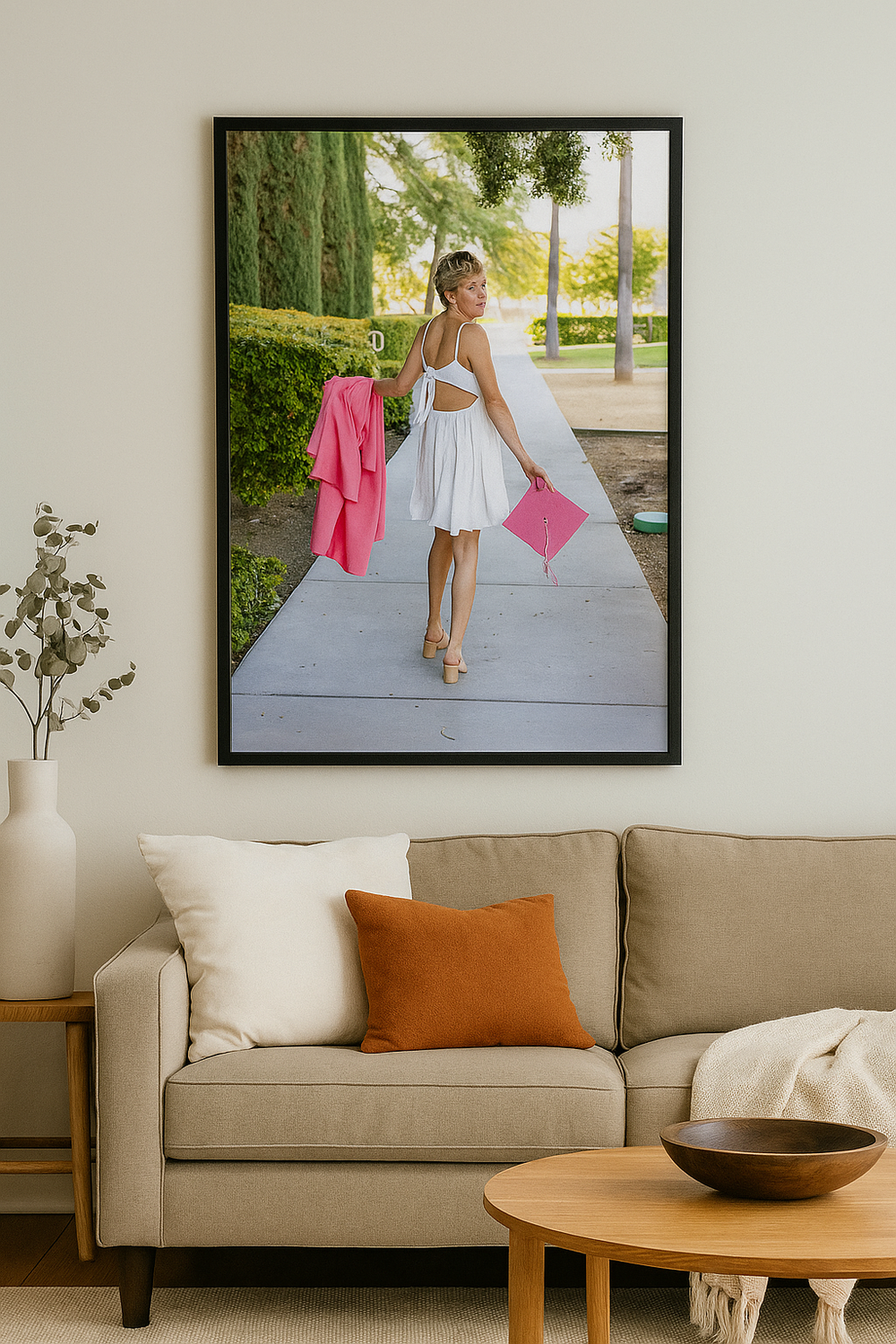A framed photo of a person in a white dress holding pink accessories hangs above a modern beige sofa with throw pillows.