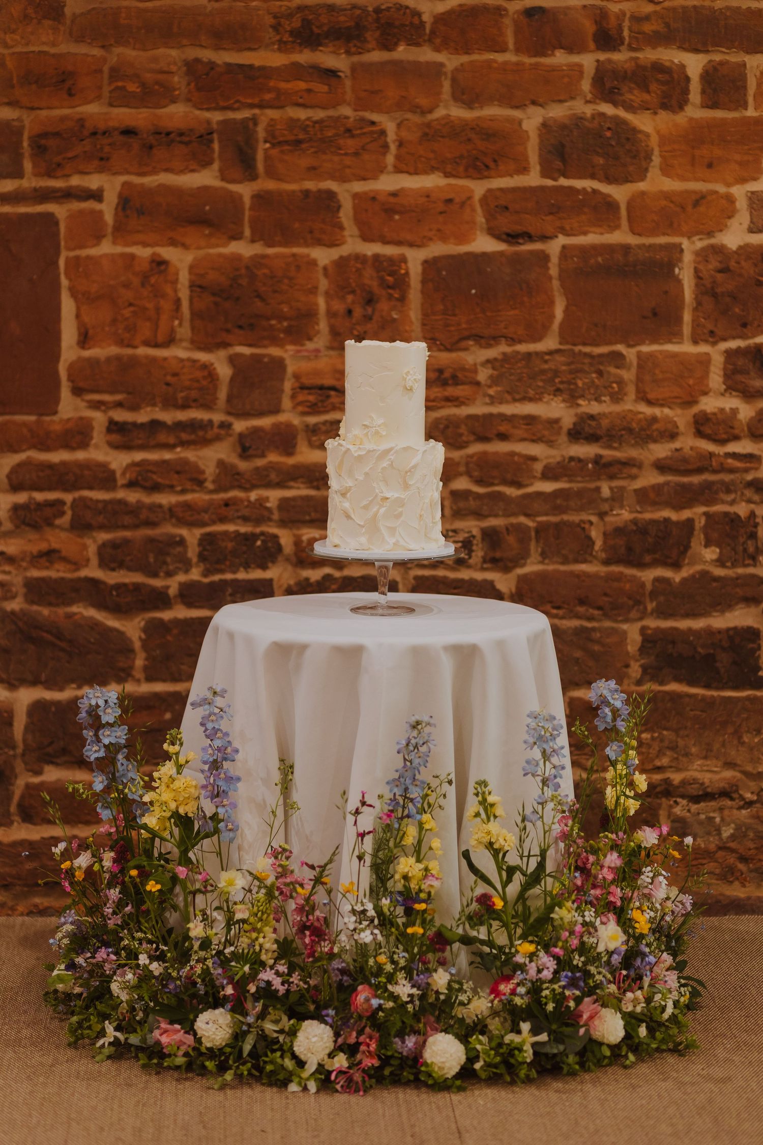 Tiered wedding cake surrounded by floral meadows against a brick wall