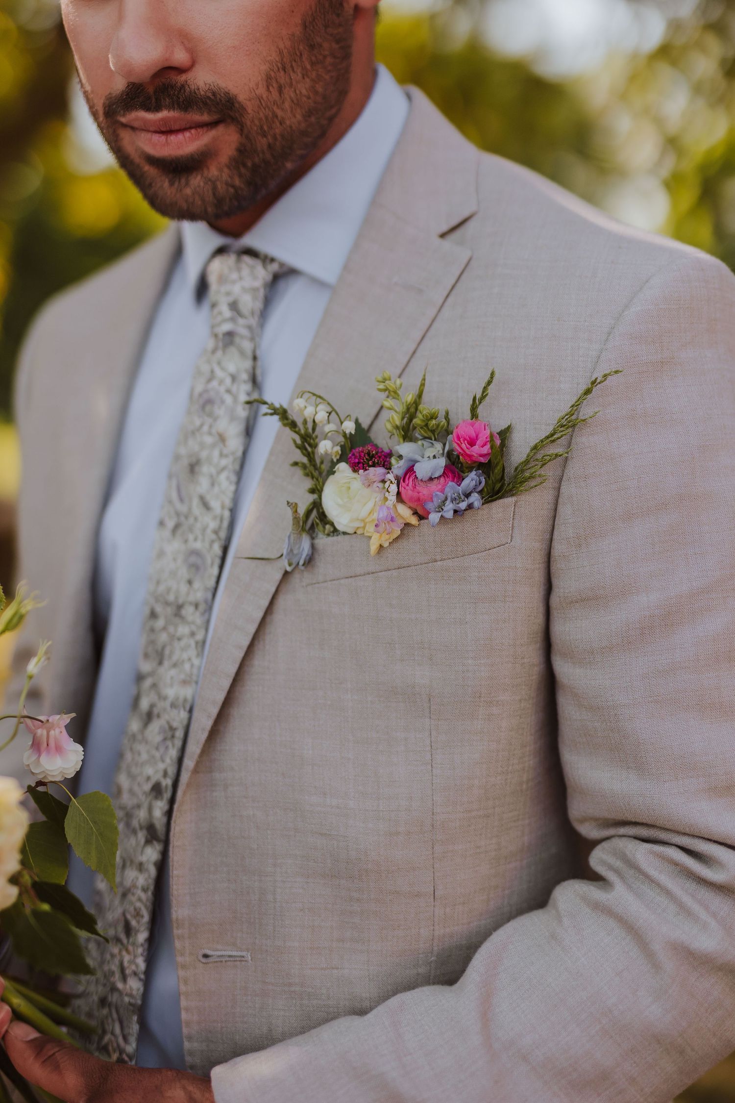 A beige suit jacket with a floral tie and pink and white boutonniere pocket square