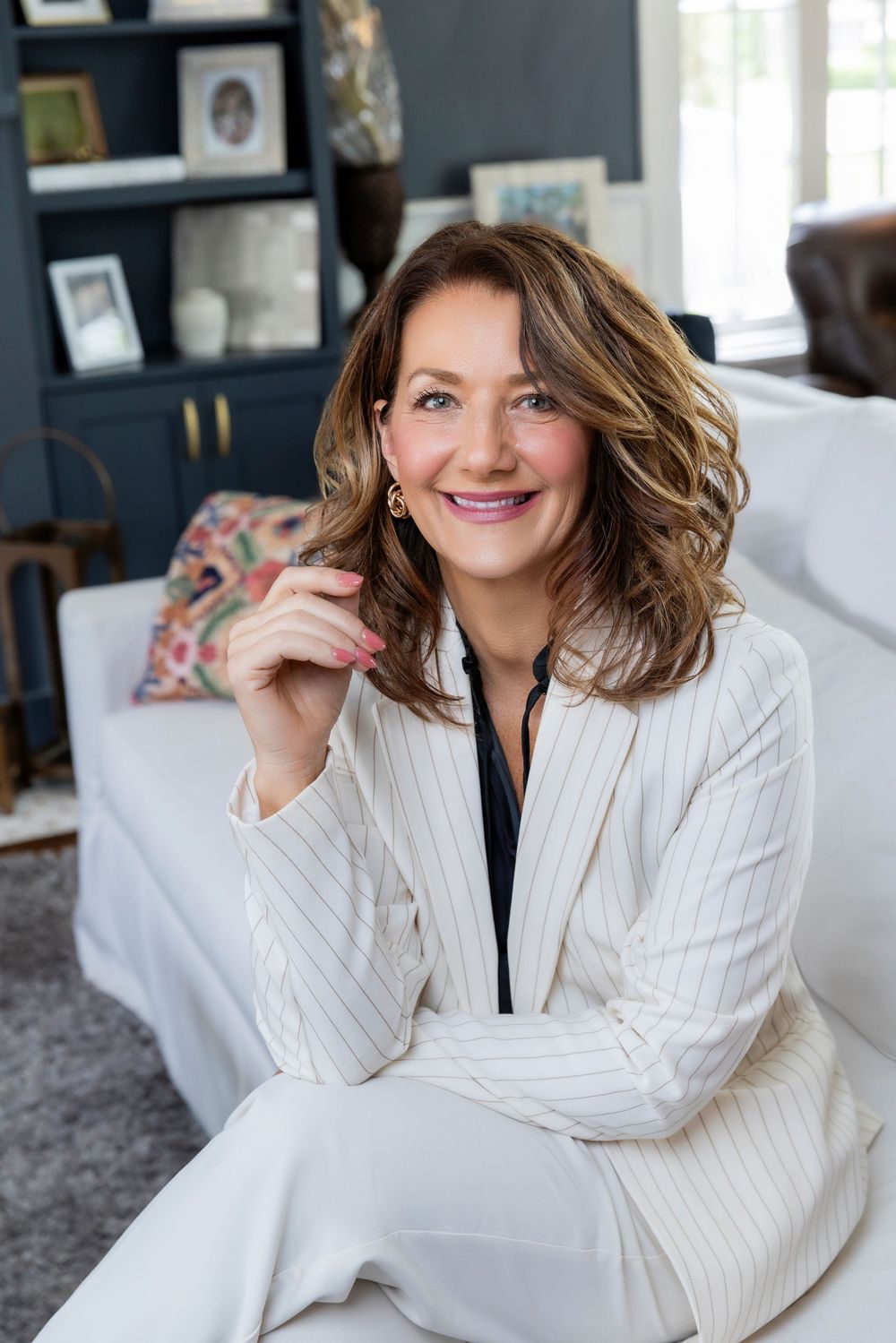 Professional portrait of someone wearing a white pinstripe blazer sitting on a couch in a modern home office setting.