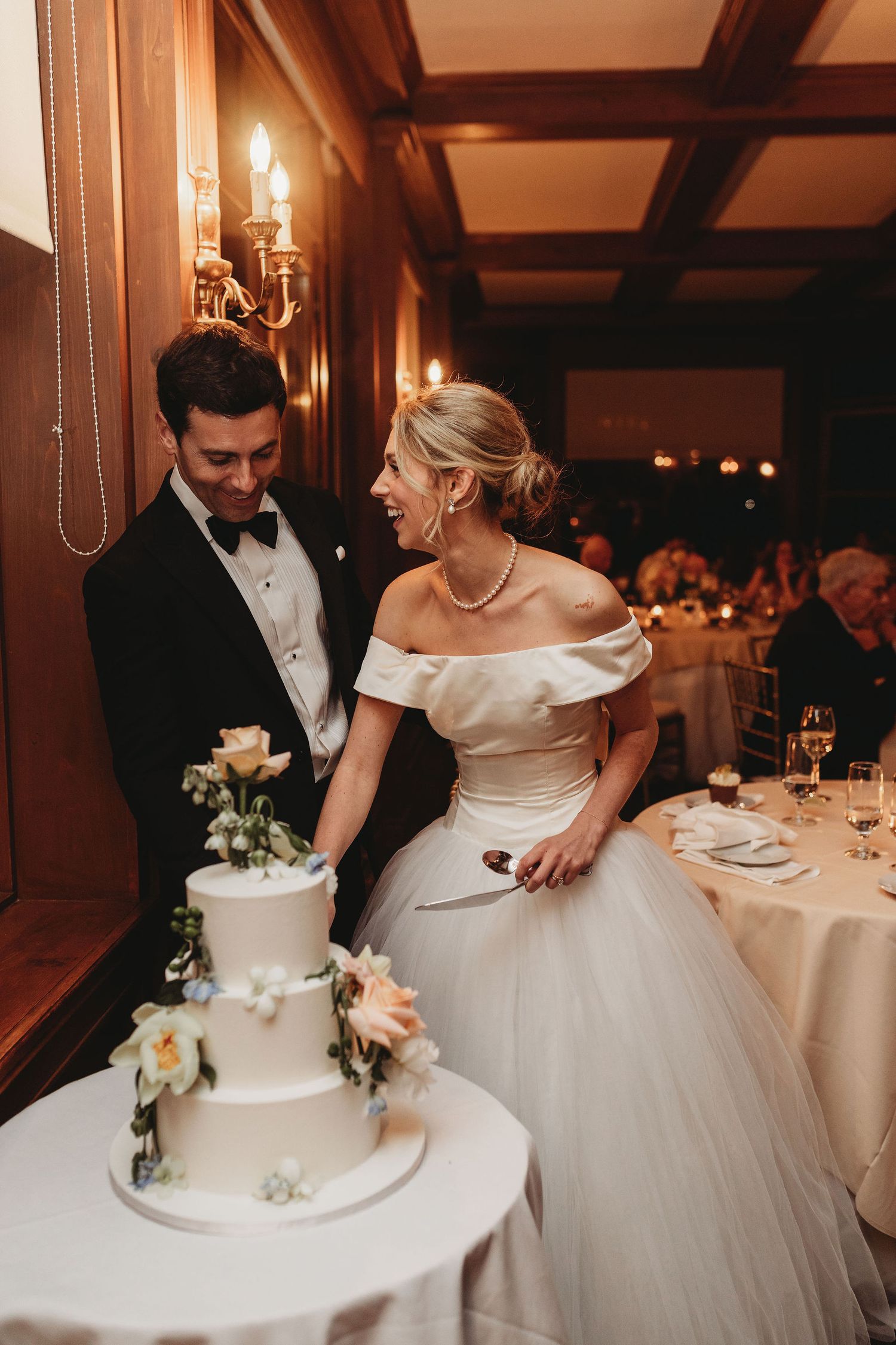 A newlywed couple cuts their elegant white wedding cake with floral decorations in a romantic candlelit reception setting.