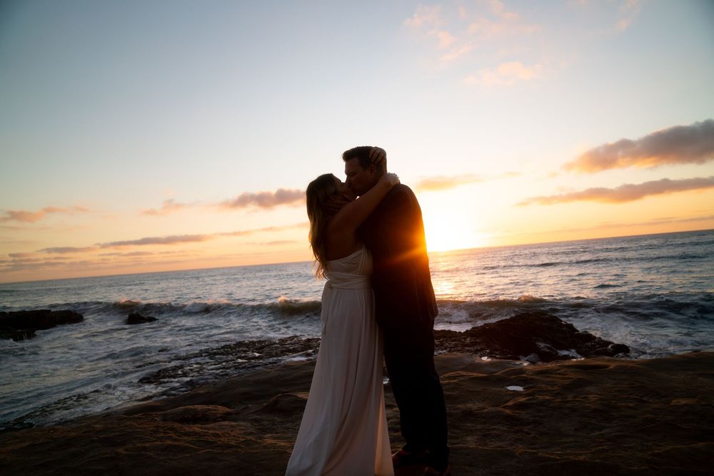 A romantic sunset silhouette of a couple embracing on a rocky beach coastline.