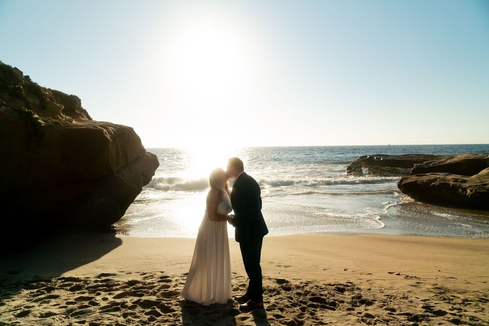Silhouetted couple embracing on sandy beach at sunset with rocky cliffs and ocean waves in background.