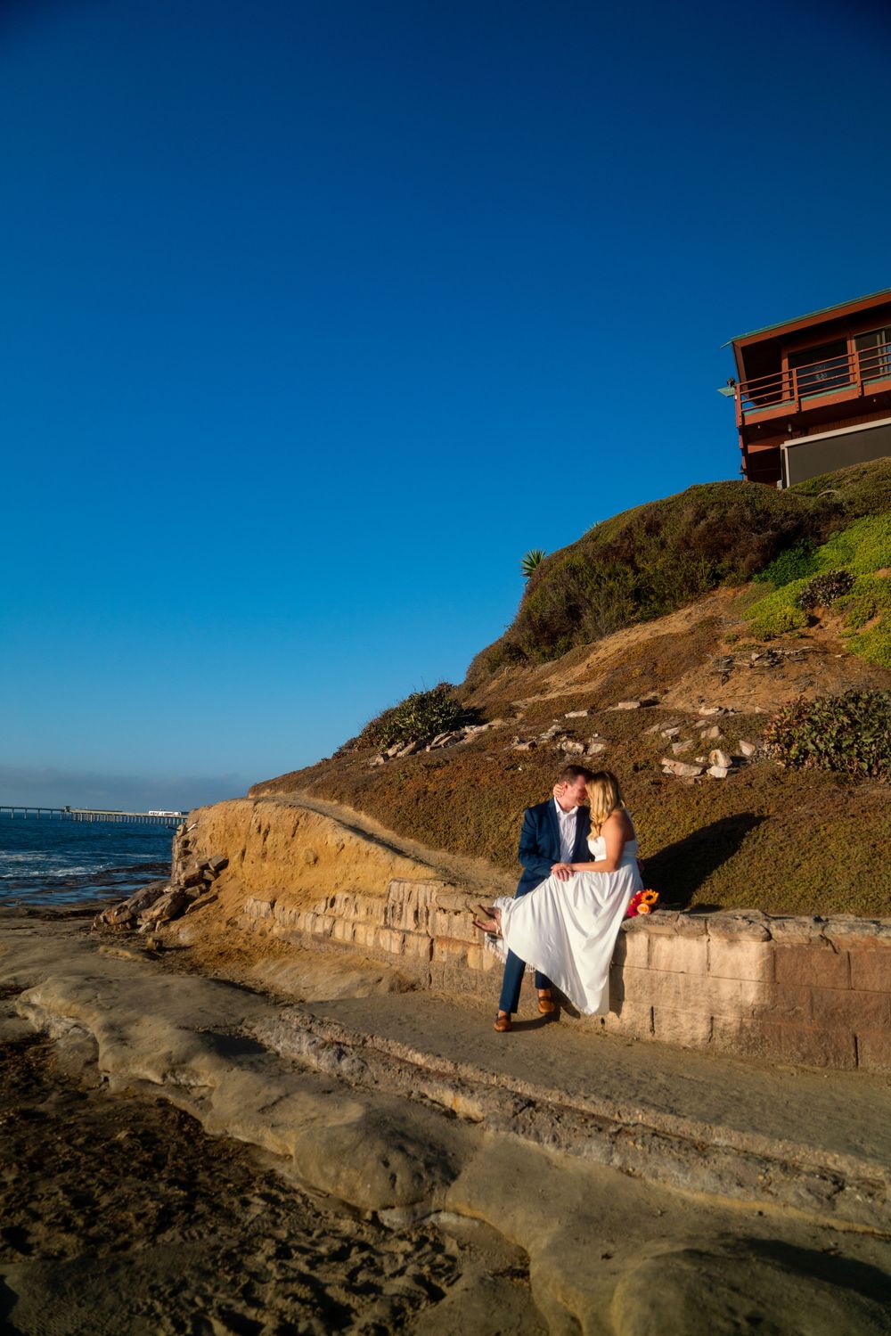 A couple sits on a coastal wall with a blue house perched on the cliff above the ocean.