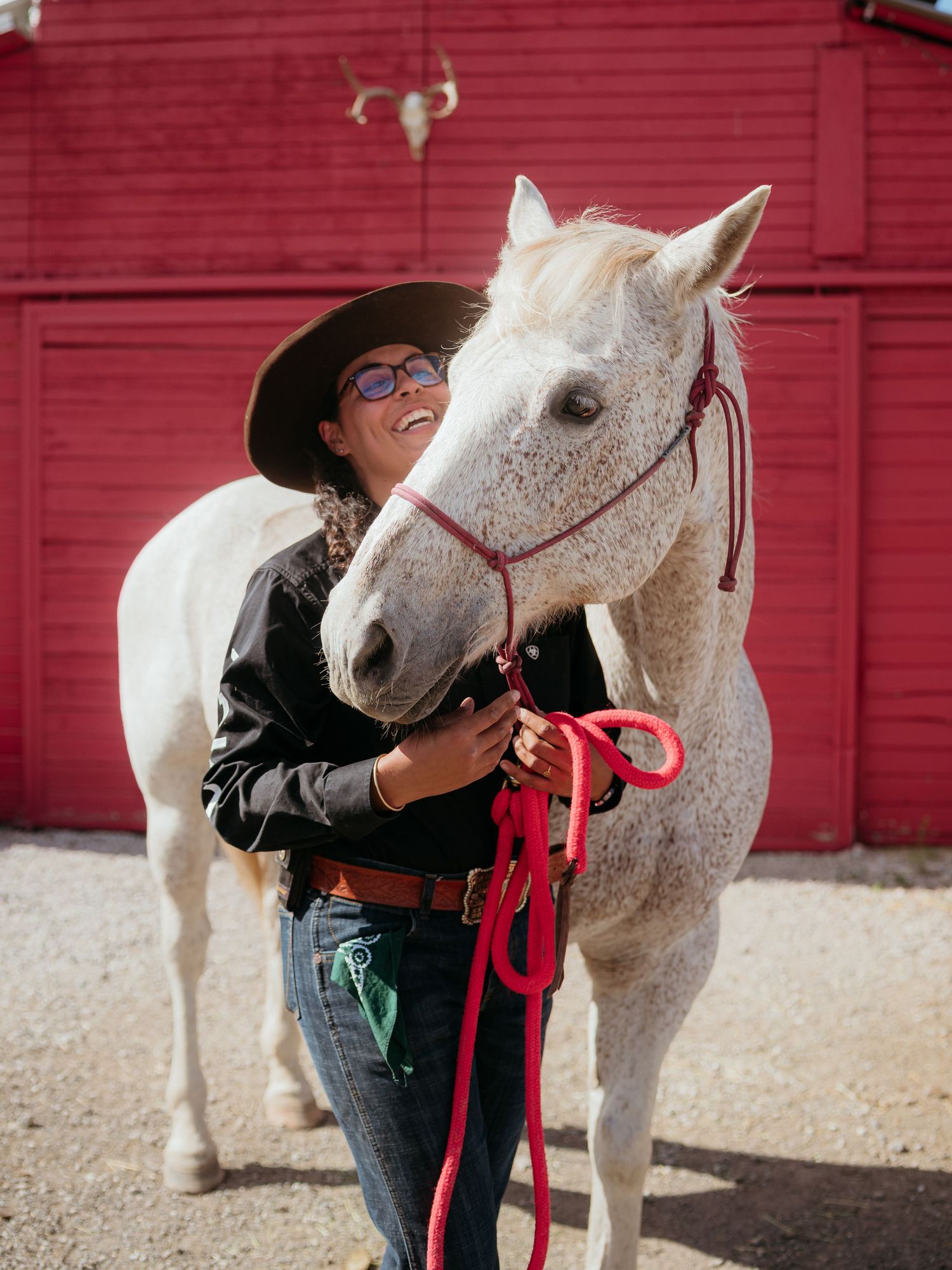 A loving moment between a grey-white horse and handler in black standing in front of a red barn on a sunny day.
