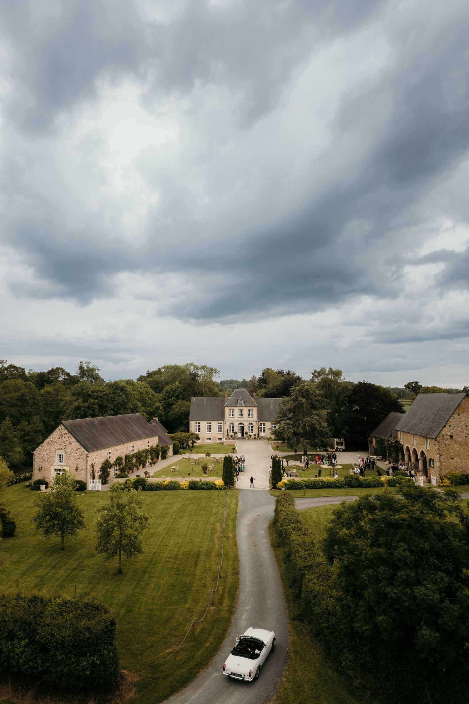 Aerial view of an elegant French country estate with manicured gardens and a vintage car driving up the winding driveway.